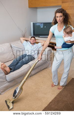 Full length portrait of young woman cleaning house while carrying baby with man relaxing on sofa