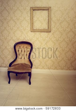 Antique upholstered chair in a wallpapered room with an empty vintage wooden picture frame hanging on the wall suitable as an interior decorating background