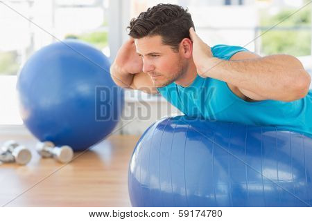 Side view of a young man exercising on fitness ball at a bright gym