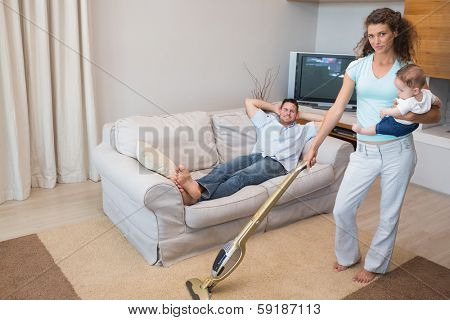 Full length portrait of woman cleaning house while carrying baby with man relaxing on sofa