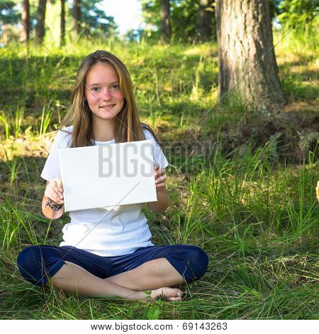 Lovely teengirl holding clean white sheet paper, in the park (banner for your message)
