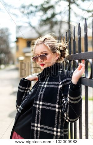 Atractive Girl In Black Coat And Sunglasses Standing Near A Wrought Iron Fence
