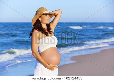 Beautiful pregnant woman on the beach with hat touching her belly