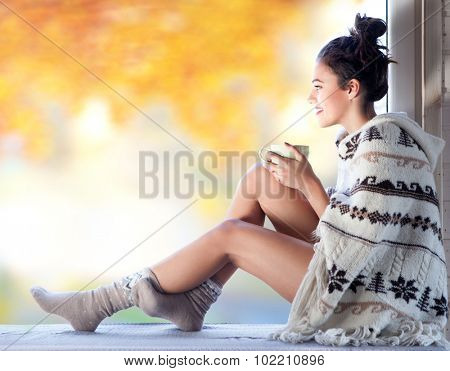 Young beautiful brunette smiling woman with cup of coffee wearing knitted nordic print poncho sitting home by the window. Blurred garden fall background. 