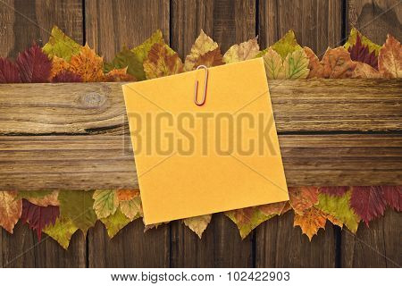 Orange adhesive note with a paperclip against autumn leaves on wood