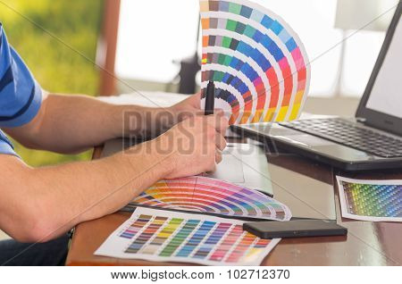 Male hands holding up palette, colormap spread out in front of laptop on working desk