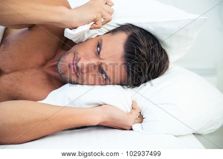 Angry man lying in the bed with pillow on head at home