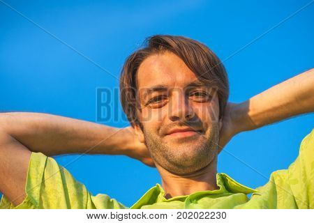 A color portrait photo of a happy smiling brunette haired man wearing a yellow lime green shirt against a blue sky backround.
