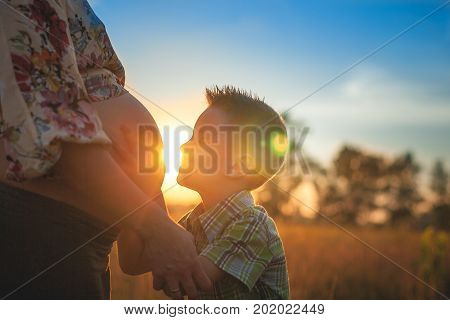 Cute boy kissing and listening his mothers pregnant mehandi pattern ornament belly on sunset nature landscape