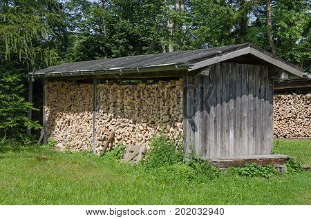 Weathered wooden shed with stacked firewood in a meadow on the edge of a forest. Simple roofed structure, used to protect firewood from moisture. Wood construction. Heuberg, Salzburg, Austria. Photo.