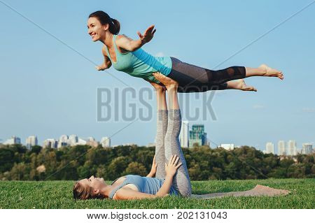 Two young beautiful Caucasian women yogi doing acro bird yoga pose. Women doing stretching workout in park outdoors at sunset. Healthy lifestyle modern activity