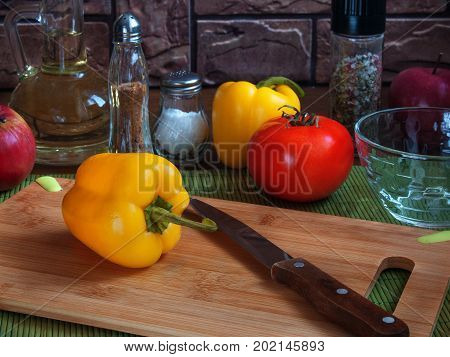 Big juicy yellow pepper on a wooden cutting board on a kitchen table with red tomatoes apples vegetable oil salt spices