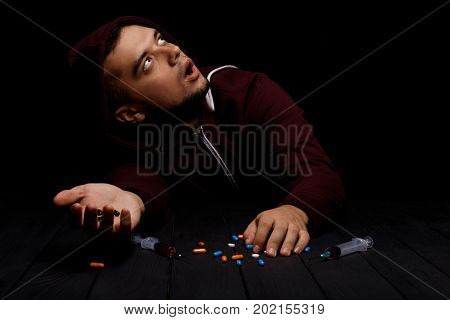 A portrait of a scared young addict man with a lot of narcotic pills and syringes full of bad red liquid on a saturated black background. An overdose of drugs. Fear, depression and narcotic concept.