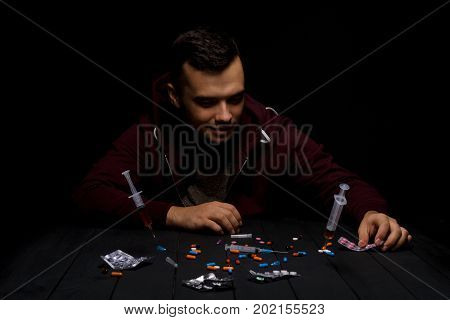 A man wearing dark red clothes sitting at a table with a lot of colorful narcotics and syringes with heroin on a saturated black background. A young guy is abusing drugs. Depression and abuse concept.
