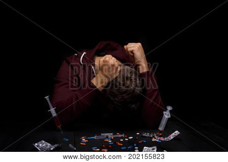 A young addict under the influence of narcotics sitting at the table on a saturated black background. A lot of pills, syringes, and drugs on a desk. An addict clings to his head with his own hands.