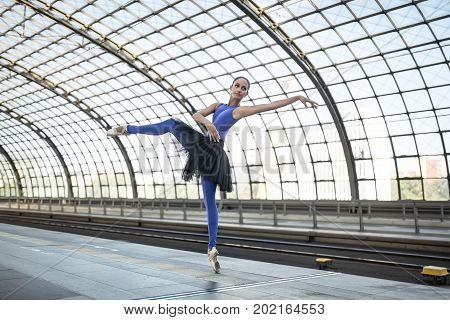 Wonderful ballerina is posing on the railway station. She stands on the right toe with outstretched to the side left arm and left leg and looks upward. Girl wears a blue dance wear with a black tutu.