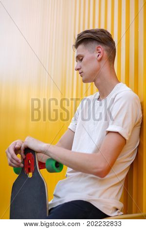 A close-up photo of a fashion young man wearing casual clothes with a modern skateboard on a bright yellow wall background. Motion, skateboarding, sports, action, and training concept. Copy space.
