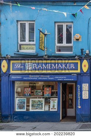Galway Ireland - August 5 2017: The dark blue facade of the small business frame maker with yellow trim open door and display window with pictures. Street scene with flags signs and wires