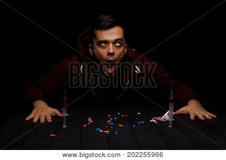 An addict sitting at the table with colorful pills and syringes on a saturated black background. An overdose of drugs. Addiction, fear, abuse and depression concept. Copy space.