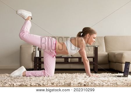 Pretty young lady in sportswear doing one knee kickbacks and watching aerobics videos on laptop, repeating exercise with online personal fitness trainer while training on carpet in living room at home
