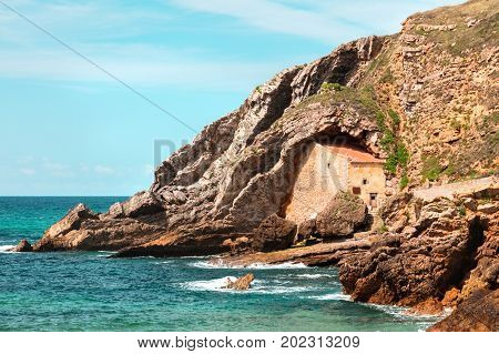 Ermita Santa Justa, a 16th century chapel built into a cove, and the blue sea and teal sky around it, with a place for text, in Cantabria