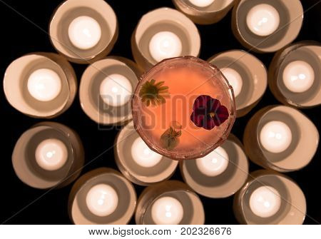 Pink cocktail garnished with flowers from above with candles. Candlelit alcoholic drink in margarita glass with pansies and daisy floating