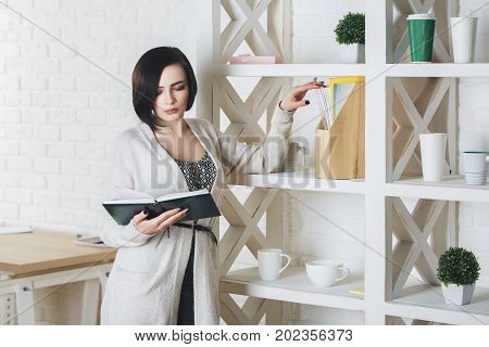 Portrait of young caucasian woman reading book in modern office interior. Knoweldge information education and leisure concept