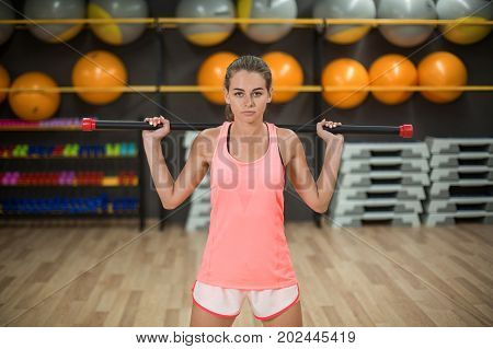 A successful, strong young woman training in pink sportswear on a blurred gym background. An attractive athlete working out with a fitness stick. Sport energy, activity, aerobics concept.