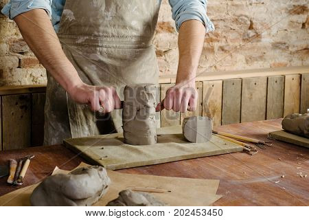 potter clay workshop ceramics art concept - male cuts raw clay with the string cutter closeup on hands of standing man dressed in an apron ceramist work over wooden table with sculpting tools set