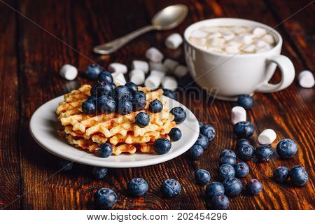 Belgian Waffles on Plate with Fresh Blueberry and Cup of Hot Cocoa with Marshmallow. Spoon and Some Berries and Marshmallows Scattered on Wooden Table.