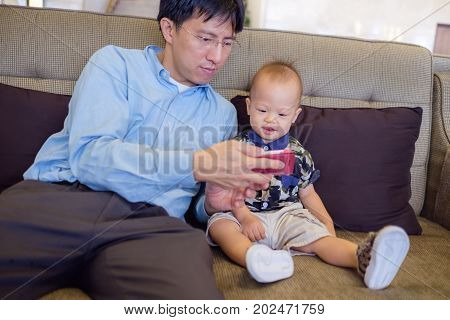 Father and son are using smartphone while spending time together Dad and Cute smiling little Asian 1 year old toddler baby boy child sitting on sofa leisure & technology & internet addiction concept