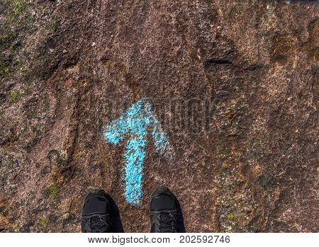 Shoes of a human standing at a blue direction arrow painted on the rock on the ground