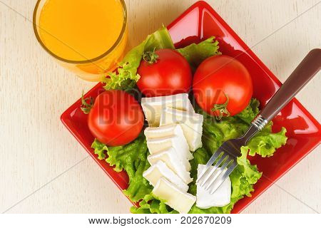 Tomatoes And Cheese Slices Lie On A Sheet Of Fresh Salad In A Red Plate. A Glass Of Orange Juice Sta