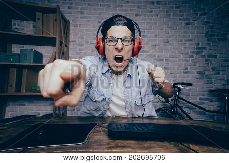 Young man in red headphones playing game at home and streaming playthrough or walkthrough video. Crazy guy is shouting because of unjustice in virtual game and pointing at the screen with one finger