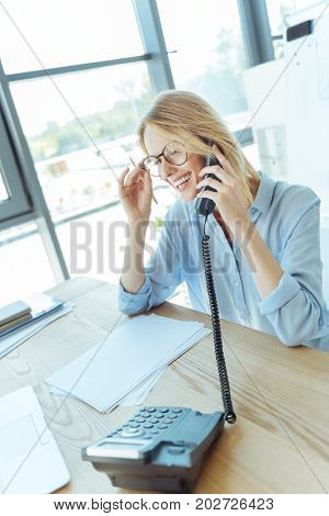 Mood-boosting conversation. Charming young woman sitting at the table and talking on the phone while smiling happily and adjusting her spectacles