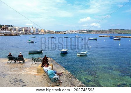 BUGIBBA, MALTA - MARCH 29, 2017 - Couples sitting on benches looking at the view across St Pauls Bay Bugibba Malta Europe, March 29, 2017.