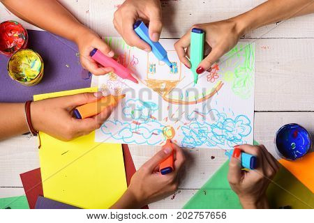 Markers in male and female hands draw on white paper. Artists wooden table with paints and colored paper. Art and artwork concept. Hands hold colorful markers and draw kids illustration top view