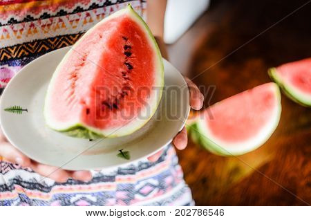 Watermelon background. Girl in a colorful summer shirt holding a plate with a large slice of watermelon against a background of a blurry table with slices of watermelon