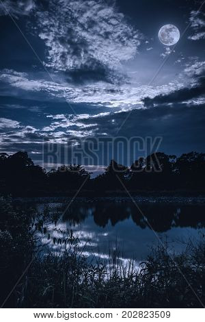 Sky With Dark Cloudy And Full Moon Above Silhouettes Of Trees And Tranquil Lake.