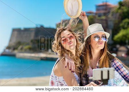 Two smiling young women sitting on a city bench making faces while taking self portraits together with a smartphone and selfie stick.