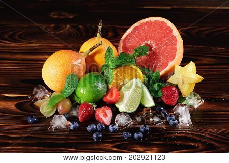 Rich assortment of many different exotic and tropical fruits: grapefruit, peaches, lime, oranges, carambola, strawberries, grapes with green mint and crushed ice on a dark wooden background.
