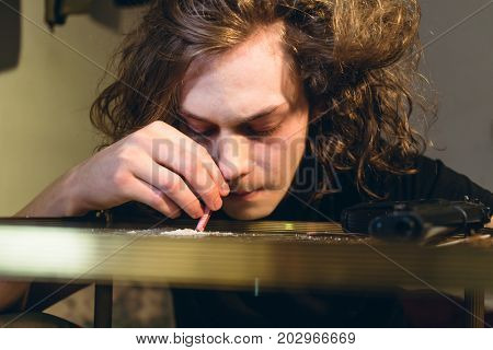 Close-up of the face of a drug addicted teenage boy holding a rolled bill while snorting cocaine from a glass table at home
