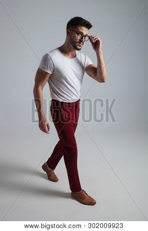 Curious man walking to the side and adjusting his glasses while wearing a white t-shirt and red pants on gray studio background