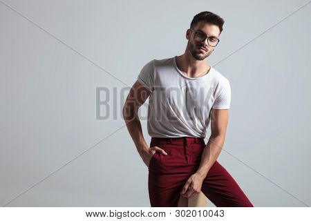 Charming guy romantically looking to the camera and holding one of his hands in his pocket while wearing glasses, a white t-shirt and red pants, sitting on gray studio background