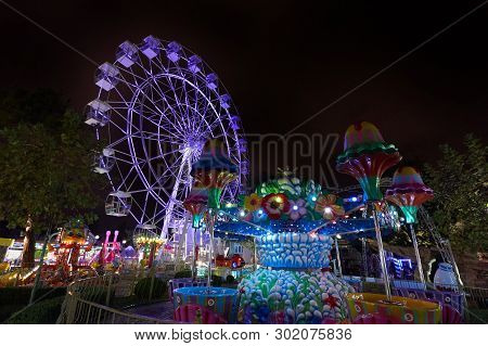 Amusement Park At Night With A Ferris Wheel And Carousels. Glowing Ferris Wheel