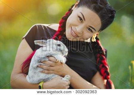 Pretty Girl Huggs Bunny. Mature Woman With Red Konekalons Hugging A Rabbit On Nature. Green Backgrou