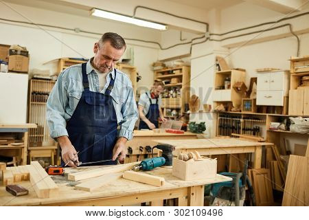 Portrait Of Mature Carpenter Working With Wood Standing At Table In Workshop, Copy Space