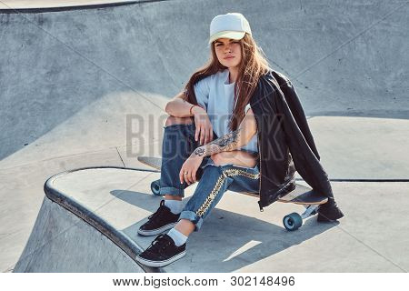 Young Attrctive Girl Is Sitting On Her Longboard At Skatepark. She Is Wearing Denim, T-shirt, Jacket