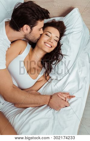 Overhead View Of Man Kissing Cheek Of Cheerful Young Woman Lying On Bed With Closed Eyes