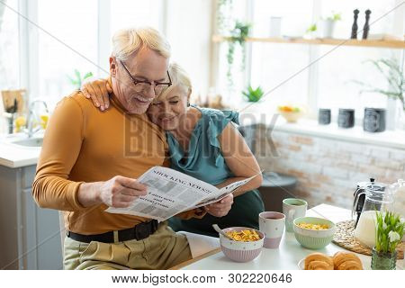 Smiling Nice-appealing Aged White-haired Married Couple Reading A Newspaper Together